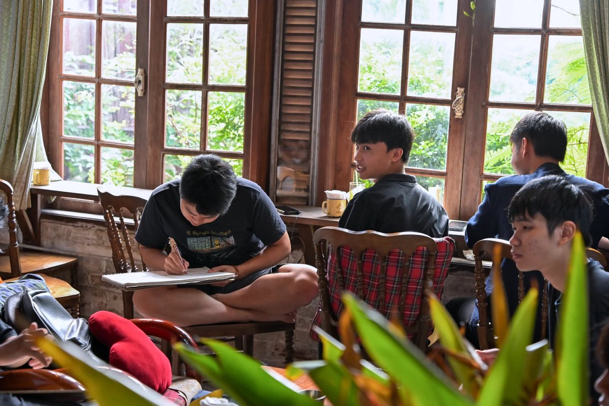 Học viên ngồi xếp bằng trên ghế gỗ, vẽ ký họa bên khung cửa sổ nhìn ra cây xanh