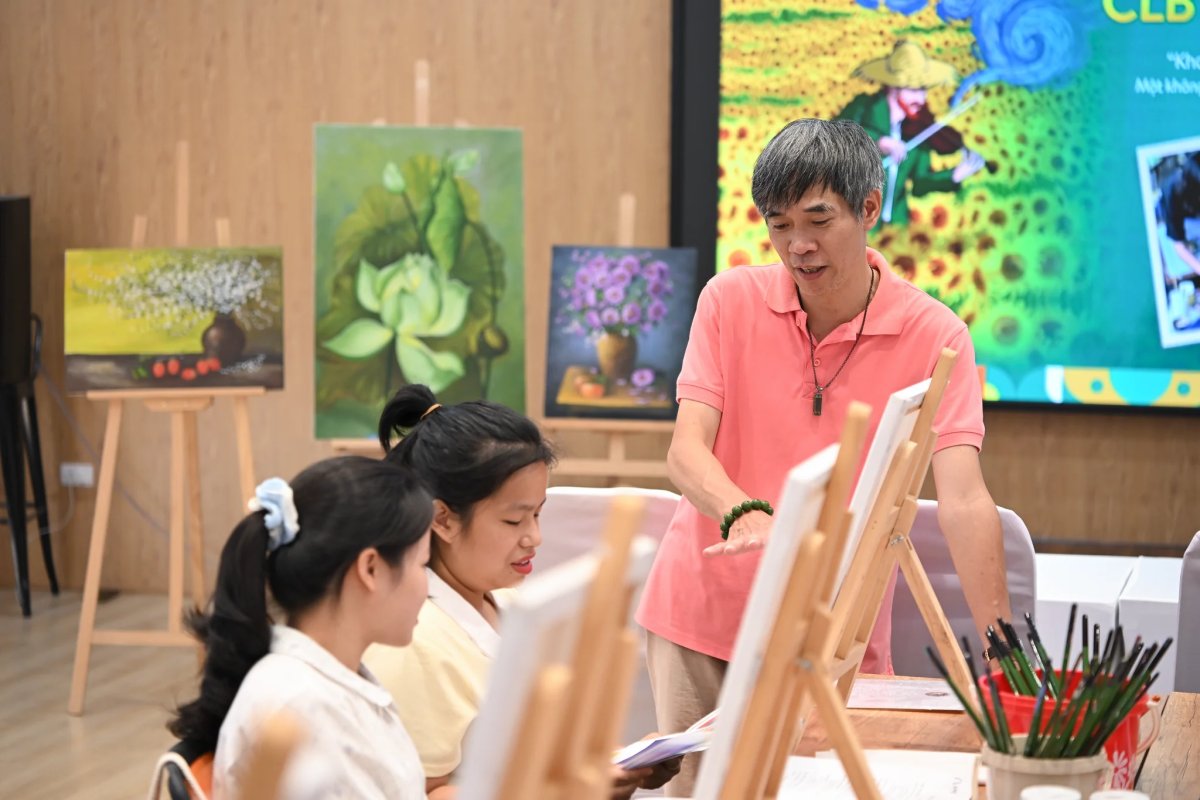 Người hướng dẫn cúi xuống bàn vẽ góp ý cho các thành viên đang vẽ trên toan